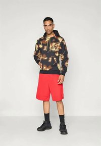 Sudadera negra con patrón de manchas coloridas, cordones amarillos y bolsillo canguro, combinada con pantalones cortos rojos y zapatillas negras.