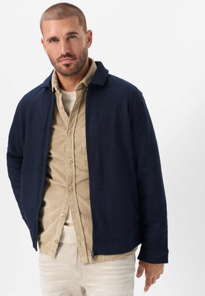 Mand med kort hår og skæg iført en navy jakke over en beige cordshirtskjorte og lyse bukser, stående foran en hvid baggrund.