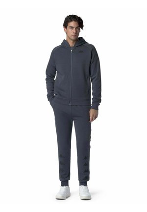 Completo da uomo con felpa con zip e pantaloni in tessuto grigio scuro. Presenta un cappuccio, polsini elasticizzati e dettagli con logo sui pantaloni. Scarpe da ginnastica bianche.