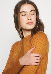 Pull à col roulé côtelé de couleur jaune moutarde profond, présentant des manches longues et une silhouette ajustée, mettant en avant un design en tricot texturé.