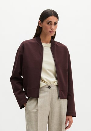 Woman with long dark hair wearing a brown zip-up jacket, cream-colored shirt, and light beige high-waisted pants against a plain white background.