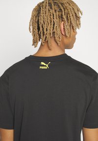 Sort T-shirt lavet af bomuld, med rund hals, korte ærmer og et gult Puma-logo trykt på den øverste del af ryggen.