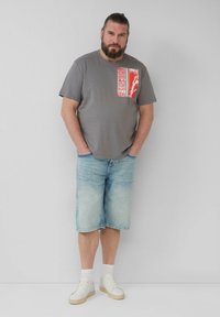 Graues Baumwoll-T-Shirt mit rotem Grafikdetail auf der Brust, kombiniert mit hellblauen Denim-Shorts und weißen Sneakers mit beigen Sohlen.