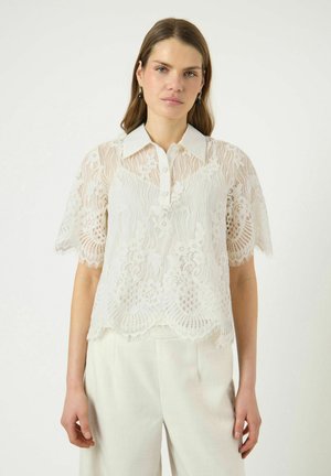 Młoda kobieta o średniej długości włosach nosi prześwitującą białą koronkową bluzkę z kołnierzykiem i krótkimi rękawami, zestawioną z białymi spodniami, na jednolitym tle.