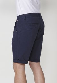 Man in een marineblauwe shorts met omgeslagen zoom en een wit shirt, staand tegen een eenvoudige lichtgrijze achtergrond.