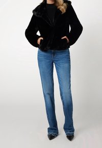 Schwarze Jacke aus Kunstpelz mit weitem Kragen, kombiniert mit hellblauen Jeans mit geradem Bein und spitzen schwarzen High Heels, mit einer glatten Textur.