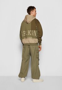 Zöld és bézs kapucnis sweatshirt, foltozott mintával és nagy felirattal. Laza, olívazöld cargo nadrággal párosítva. Fehér sportcipő.