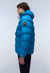 Gestikte felblauwe jas met capuchon, met een logopatch op de mouw en een gestructureerde afwerking, gedragen over zwarte broek.