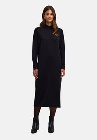 Robe noire à manches longues avec un col haut, fabriquée dans un tissu doux. Le modèle porte des bottines noires, se tenant devant un fond uni.