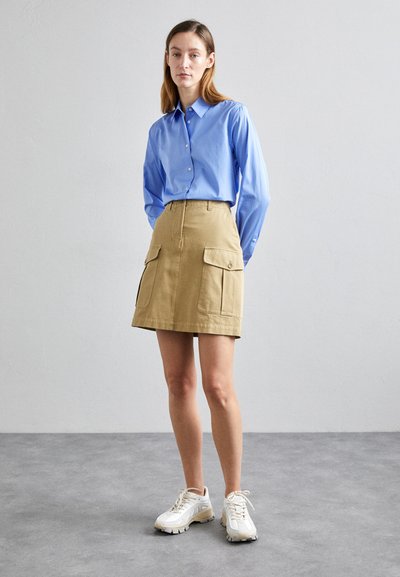 Camicia azzurra con bottoni e maniche lunghe, gonna cargo beige con due tasche frontali e sneakers bianche, su uno sfondo neutro.