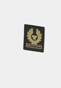 Rektangulær tekstilpatch i svart med et gull-emblem som har vinger og en logo, merket "BELSTAFF" nederst. Teksturert overflate.