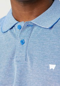 Polo shirt bleu clair avec un tissage texturé, présentant un col et des boutons bleus contrastants, ainsi qu'un logo blanc sur le côté gauche.