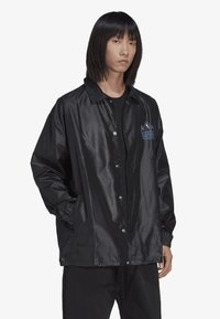 Veste de coach en satin noir avec des boutons-pression, des poches latérales et un logo de couronne bleue brodé sur la poitrine gauche. Texture lisse, coupe décontractée.