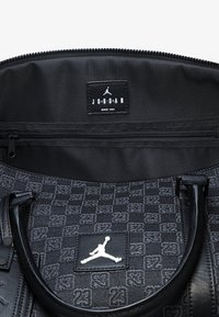 Μαύρη αθλητική τσάντα με υφασμάτινη υφή, λογότυπο "23", δερμάτινες λεπτομέρειες, σήμα Jordan και εσωτερική θήκη με φερμουάρ.