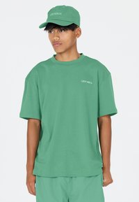 Grønt oversized t-shirt med rund hals og korte ærmer, parret med en matchende grøn baseballcap med teksten "LES'ARCS".