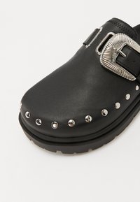 Black synthetic clog with a rounded toe, silver buckle detail, and studded accents along the sole. Textured surface and thick rubber outsole.