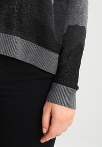 Šedý pletený svetr s přechodem barev a různými texturami, včetně lesklých a matných částí, s žebrovanými manžetami a volným střihem.