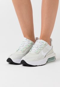 White athletic shoes with green accents, synthetic upper, mesh panels, round laces, and a visible air cushioning unit in the sole.