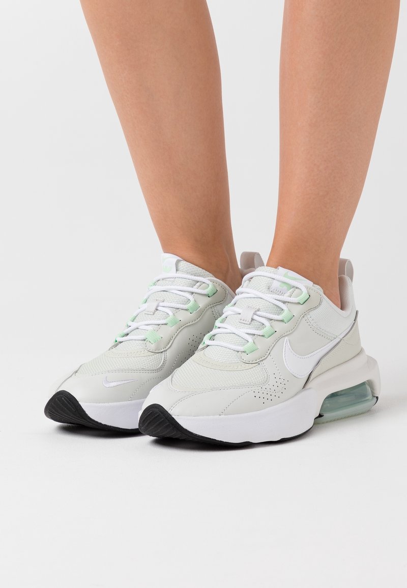 White athletic shoes with green accents, synthetic upper, mesh panels, round laces, and a visible air cushioning unit in the sole.