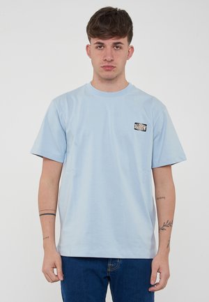 T-shirt in cotone azzurro chiaro con maniche corte, collo a giro e una piccola etichetta grafica nera sul lato sinistro del petto. Il tessuto sembra liscio.