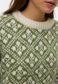Pull en tricot avec un col rond crème. Présente un motif géométrique vert et blanc, la texture est épaisse et duveteuse.