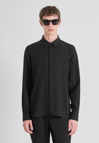Camisa negra de manga larga con cuello abotonado, textura suave y corte holgado. Modelo lleva gafas de sol negras para un look monocromático.