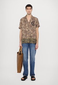 Camisa de manga corta con estampado de leopardo y logo negro, combinada con jeans azul claro y sandalias negras; sosteniendo un bolso tote marrón brillante.