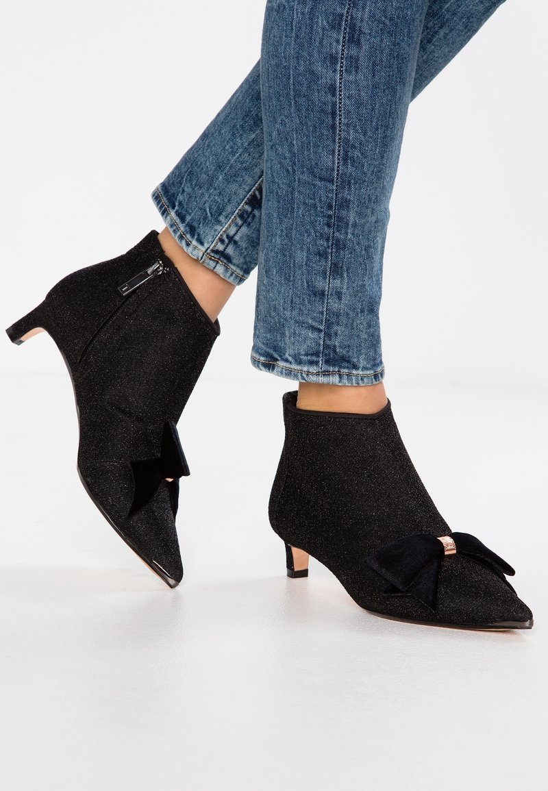 Bottines à paillettes noires avec un bout pointu, dotées d'un accent de nœud noir et d'un petit talon. Tissu texturé et détail de fermeture éclair sur le côté.