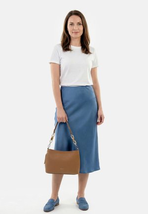 Woman in white short-sleeve shirt, blue midi skirt, and blue loafers holding a brown handbag against a plain white background.