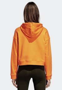 Oranžová zkrácená mikina s kapucí na šňůrku, měkká textura a volný střih, v kombinaci s těsnými tmavými legínami.