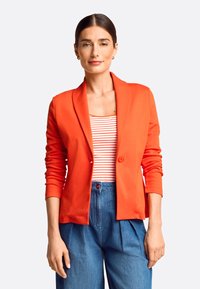 Roter Blazer mit einem einzigen Knopf, aus weichem Stoff und taillierter Passform. Getragen über einem gestreiften Tanktop und kombiniert mit hochgeschnittenen blauen Hosen.