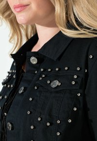 Svart denimjacka med silverfärgade dekorativa nitar, två bröstfickor och silvriga knappar. Har en klassisk krage och strukturerad tyg.