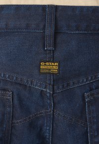 Tamno plavi traperice od denima sadrže G-Star Originals RAW etiketu. Dizajn uključuje vidljive šavove i glatku teksturu.