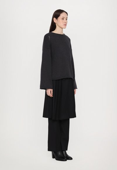 Tmavě šedý svetr s nadměrnými rukávy, v kombinaci s černou plisovanou sukní a elegantními černými kalhotami. Modelka má na sobě černé boty na podpatku.