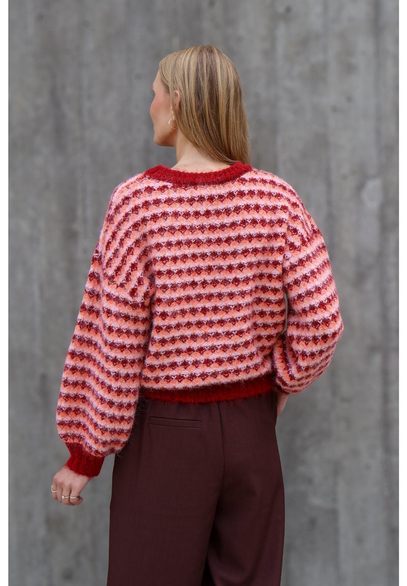 Femme aux longs cheveux blonds porte un pull en tricot rouge et rose à motifs et un pantalon rouge foncé, debout contre un mur en béton gris.