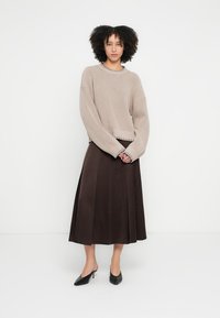 Bézs kötött pulóver texturált felülettel és széles ujjal, barna A-vonalú szoknyával párosítva. A megjelenést fekete hegyes orrú cipő teszi teljessé.