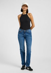 Svart ribbad halterneck-topp i kombination med blå raka jeans. Modellen bär svarta sneakers med tre vita ränder och en vit keps.