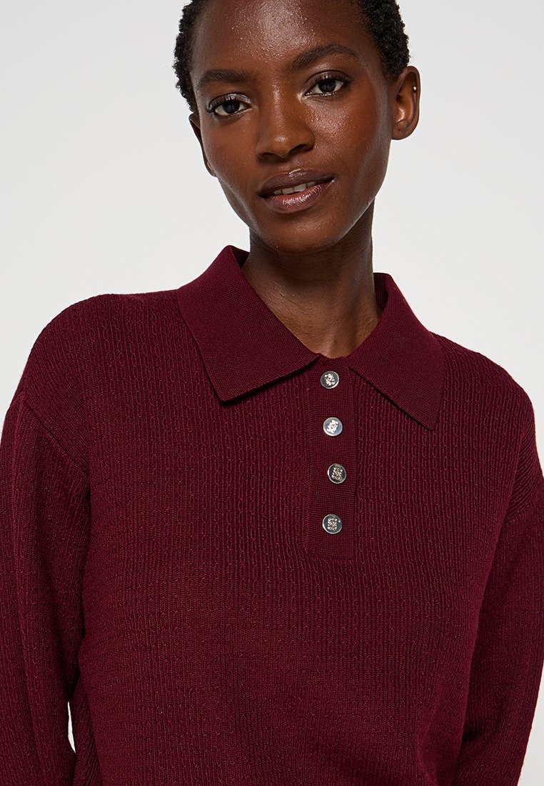 Polo shirt lavorato in maglia color borgogna con un tessuto texturizzato, caratterizzato da un colletto aperto e cinque bottoni metallici sulla parte anteriore.