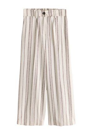 Vrouw loopt, draagt een beige en wit vertical gestreepte mouwloze vest en bijpassende wijde broek met zwarte sandalen tegen een gestructureerde muur.