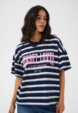Kobieta z falującymi ciemnymi włosami, ubrana w granatowo-jasnoniebieski pasiasty t-shirt z różowym napisem i ciemny naszyjnik z koralików.