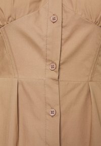 Jasnobrązowy materiał z pionowymi szwami i trzema okrągłymi, dopasowanymi guzikami na środku przodu odzieży.