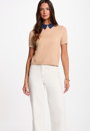 Mujer de pie con suéter beige de manga corta con cuello azul oscuro y pantalones blancos de talle alto, posando sobre fondo blanco.