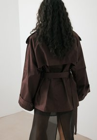 Brązowy płaszcz trench wykonany z materiału odpornego na wodę, z paskiem w talii, szerokimi rękawami i warstwowym krojem, na przezroczystej czarnej tkaninie.