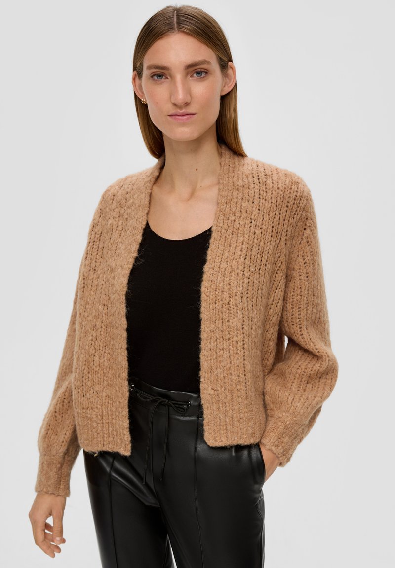 Strickjacke in sanftem Beige mit langen Ärmeln, offener Front und geripptem Muster. Getragen über einem schwarzen Oberteil und kombiniert mit schwarzen Hosen.