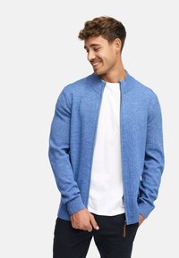 Cardigan zippé bleu en maille texturée. Il présente un col montant et des poignets côtelés, porté par-dessus une chemise blanche.