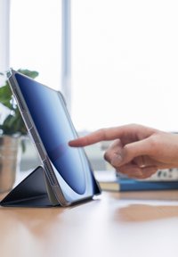 Eine Hand zeigt auf den aufrecht stehenden Tablet-Bildschirm, der auf einem Schreibtisch steht, im Hintergrund sind Bücher und eine Pflanze zu sehen.