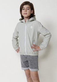 Giacca a zip grigio chiaro con cappuccio, polsini ed orlo elasticizzati. Presenta un logo bianco sul petto e tasche laterali. Indossata con pantaloni corti grigi.