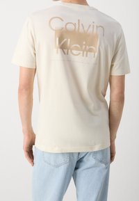 Camiseta de algodón crema con un gran logo "Calvin Klein" en color marrón claro en la parte posterior, con un corte relajado y mangas cortas.