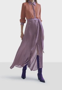 El vestido maxi presenta un patrón de cuadros rojos y morados, con un cuello tipo camisa, mangas largas y una cintura atada. Se combina con botas moradas con estampado.