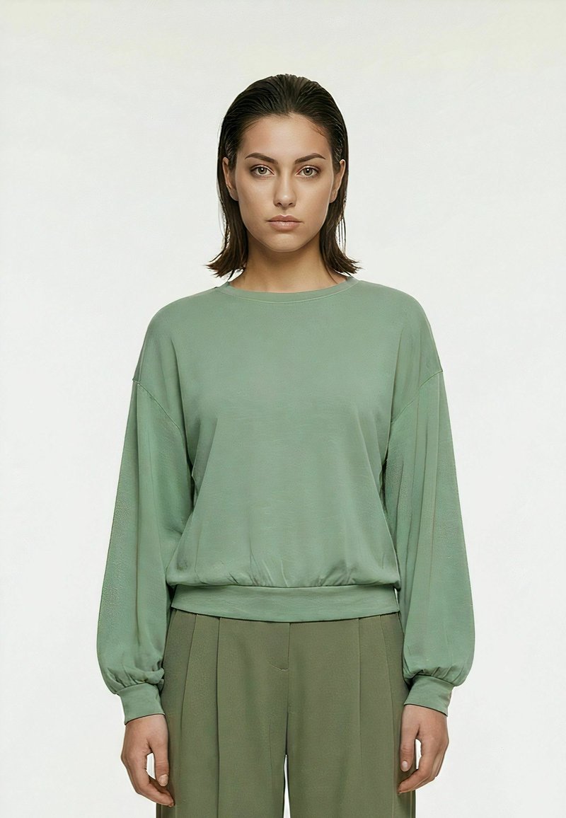 Femme aux cheveux raides et foncés portant un haut ample à manches longues vert sauge et un pantalon plissé vert olive assorti, debout devant un fond uni.